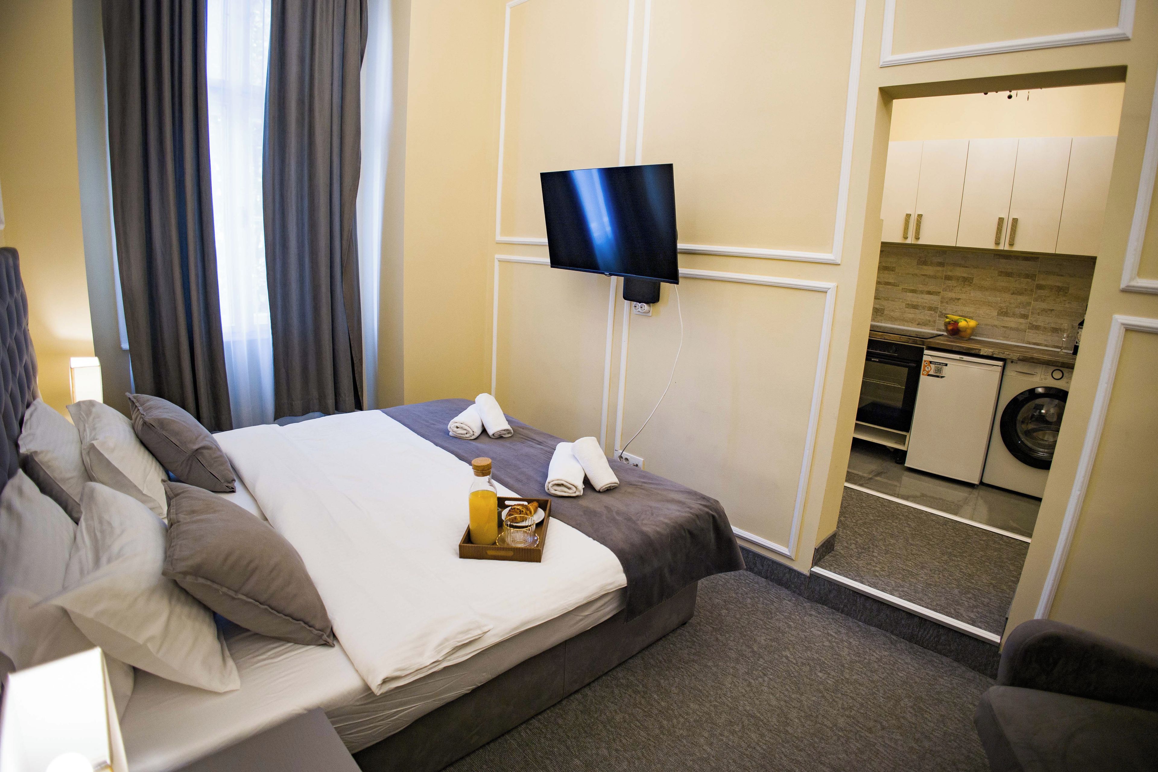 A hotel room with a bed, a TV mounted on the wall, a small kitchenette, and a bathroom visible through an open door.