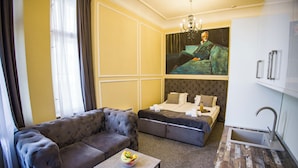 Een moderne hotelkamer met een hoekbank, een tafeltje met fruit, een bed met kussens, een schilderij aan de muur en een wastafel.