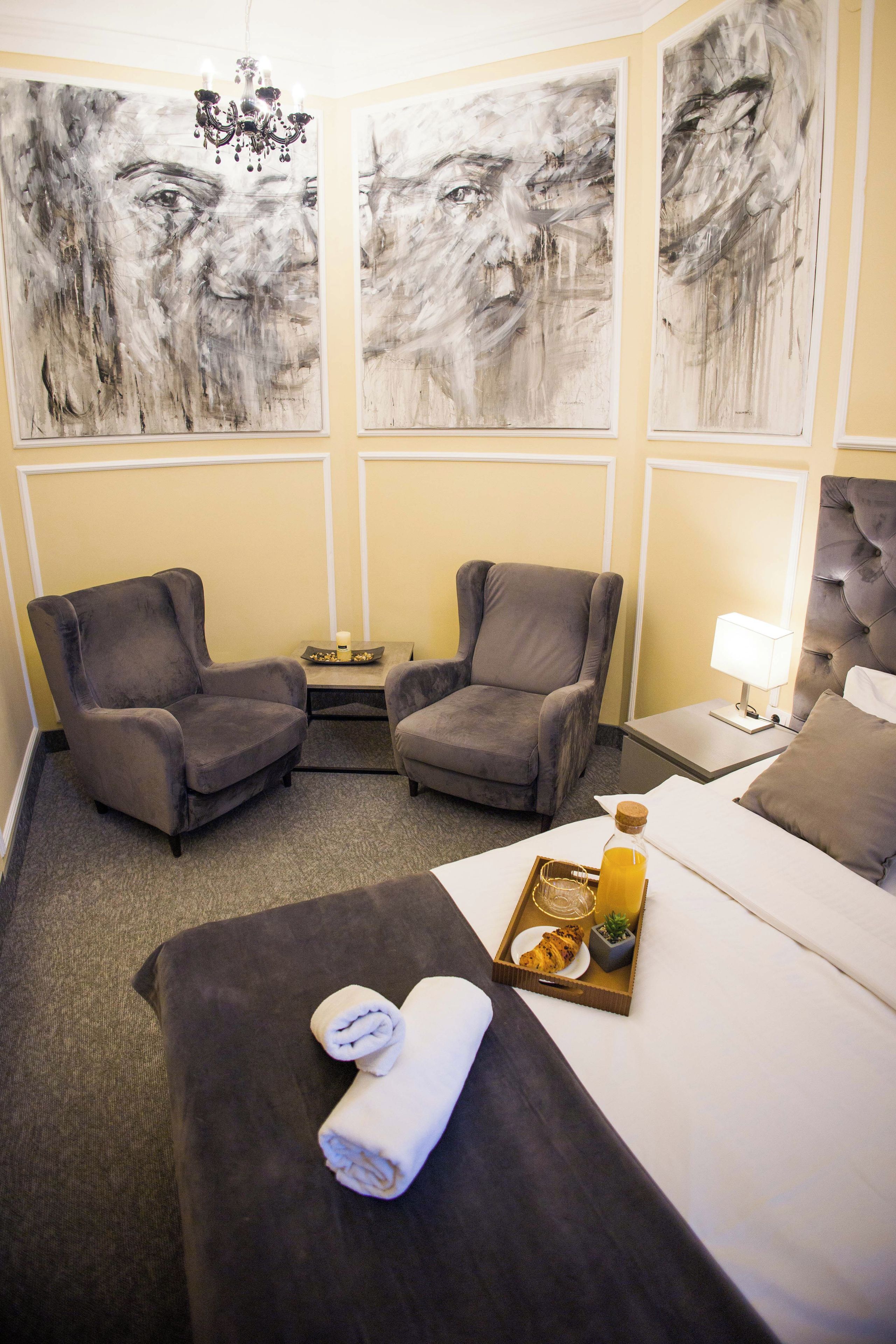 Hotellihuone, jossa on sänky, kaksi nojatuolia, yöpöytä lampun kanssa ja kolme suurta abstraktia maalausta seinällä.
