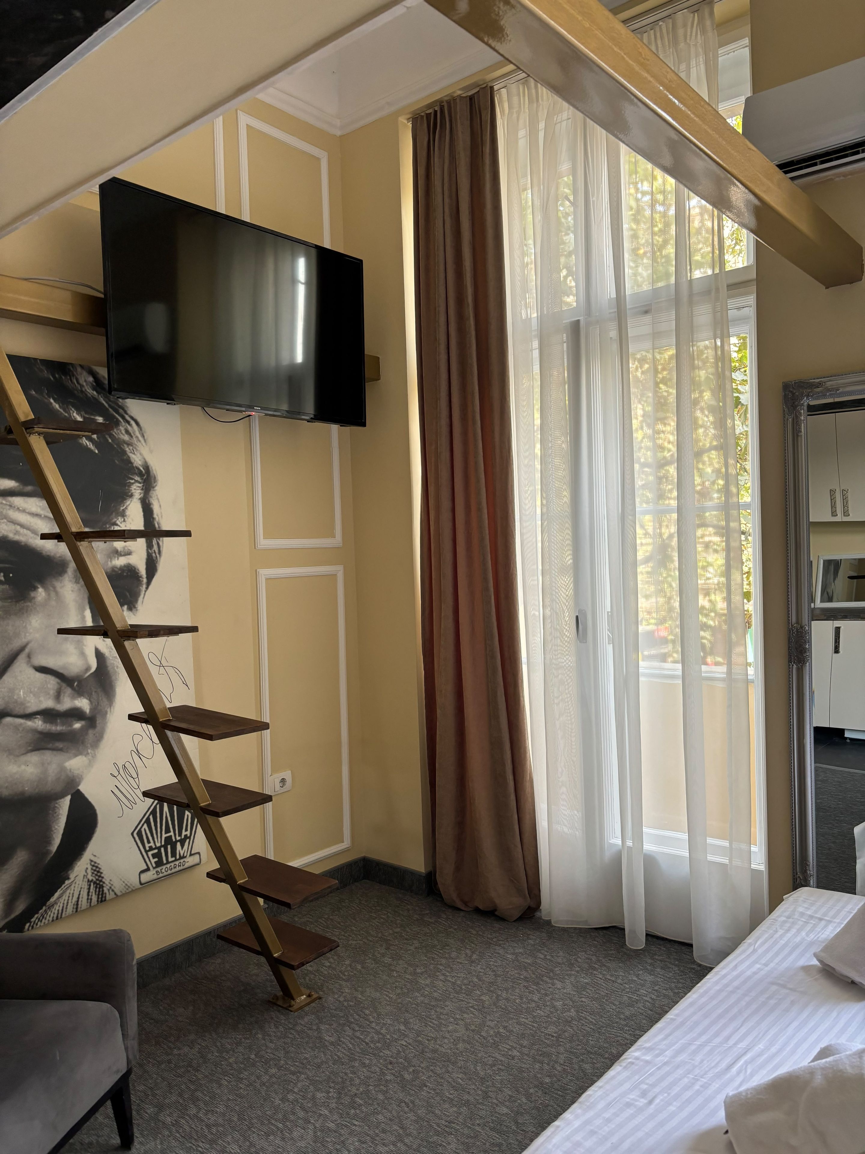 Une pièce comprenant un lit, un téléviseur fixé au mur, un escalier et une fenêtre avec des rideaux.