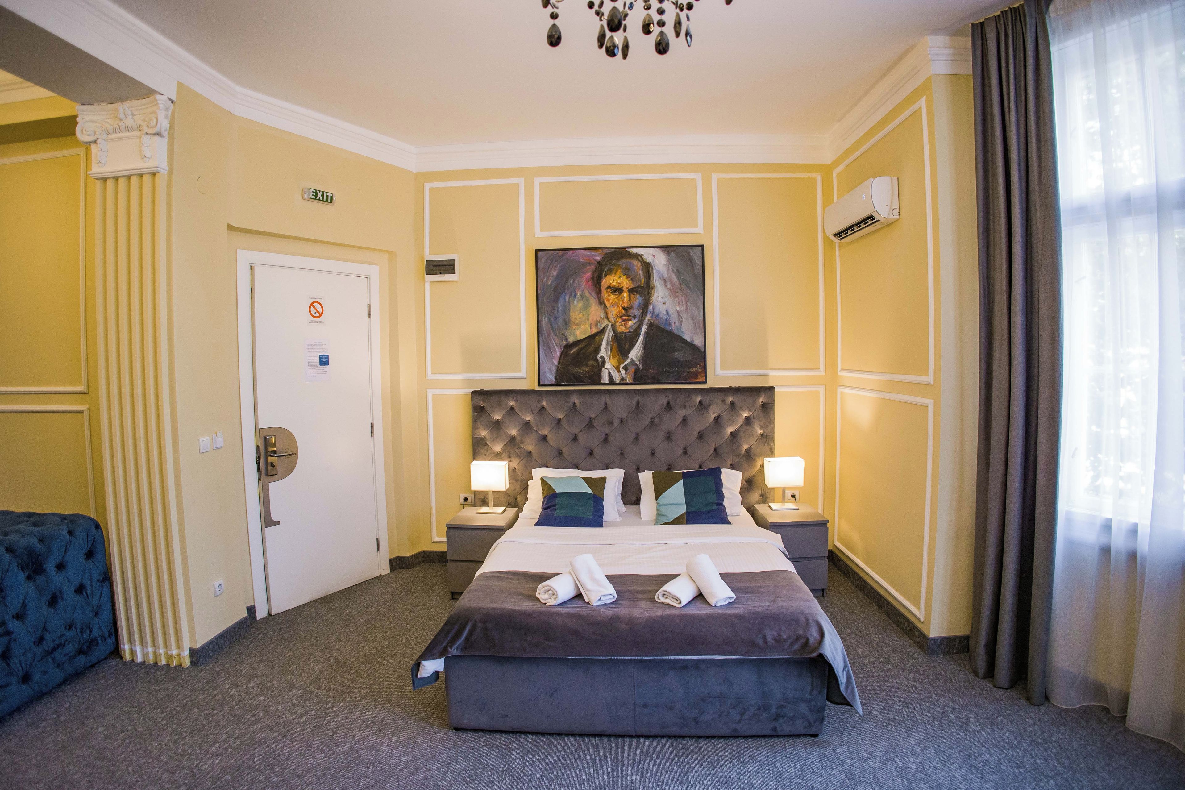 A hotel room with a large bed, two bedside tables with lamps, a painting on the wall, and a door with an exit sign.