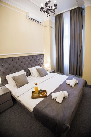 A hotel room with a large bed, a bedside table, a nightstand, a chandelier, and a window with curtains. - The Velvet suites (Beograd)