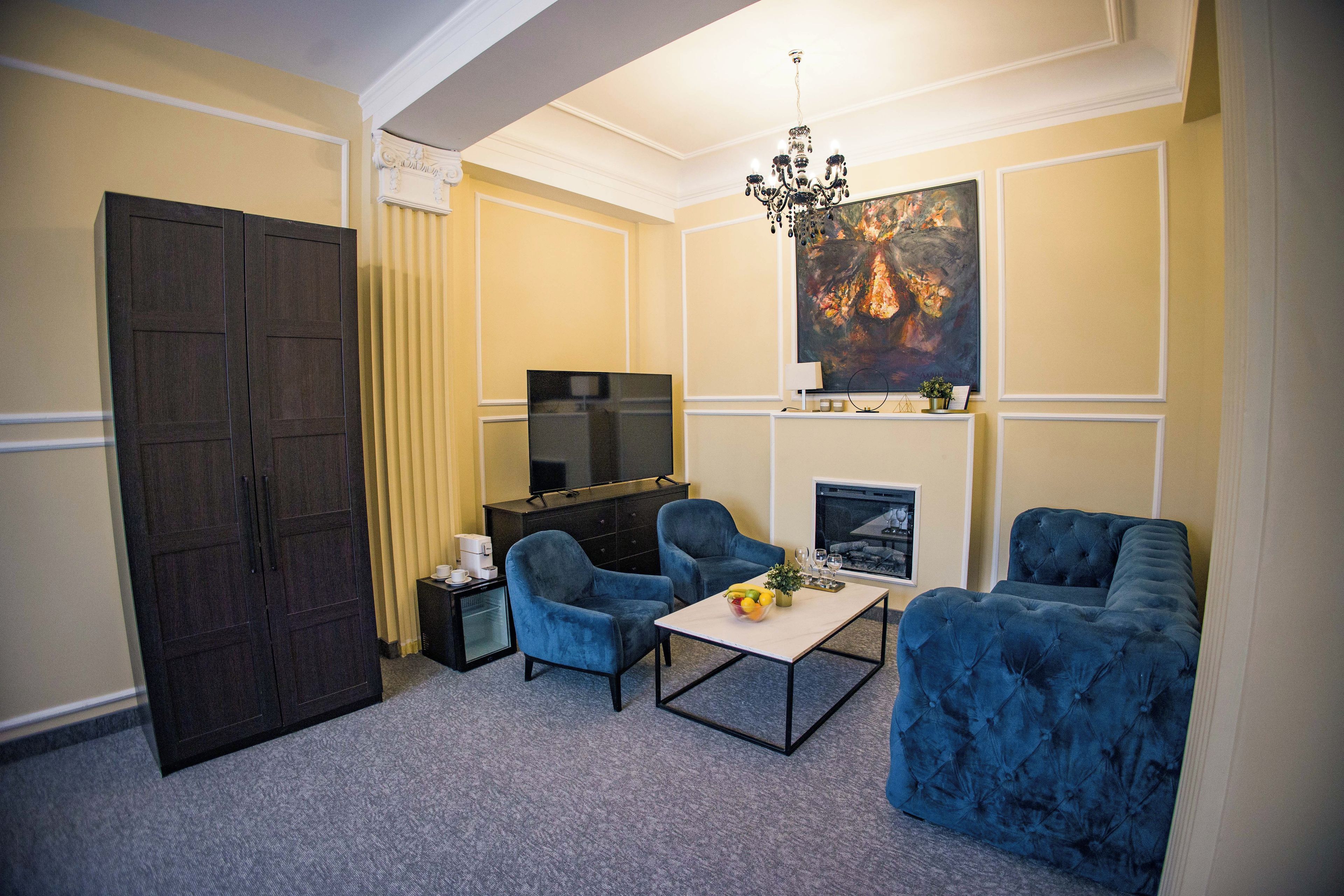 Sala de estar com porta de madeira escura, duas poltronas azuis, uma mesinha com uma tigela de frutas, televisão e lareira.
