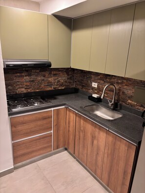 A kitchen with a gas stove, wooden cabinets, a black countertop, and a stone backsplash. - Charming Apartment in Piedra Pintada | 2 Bedrooms, Pool & Mountain Views (Ibagué)