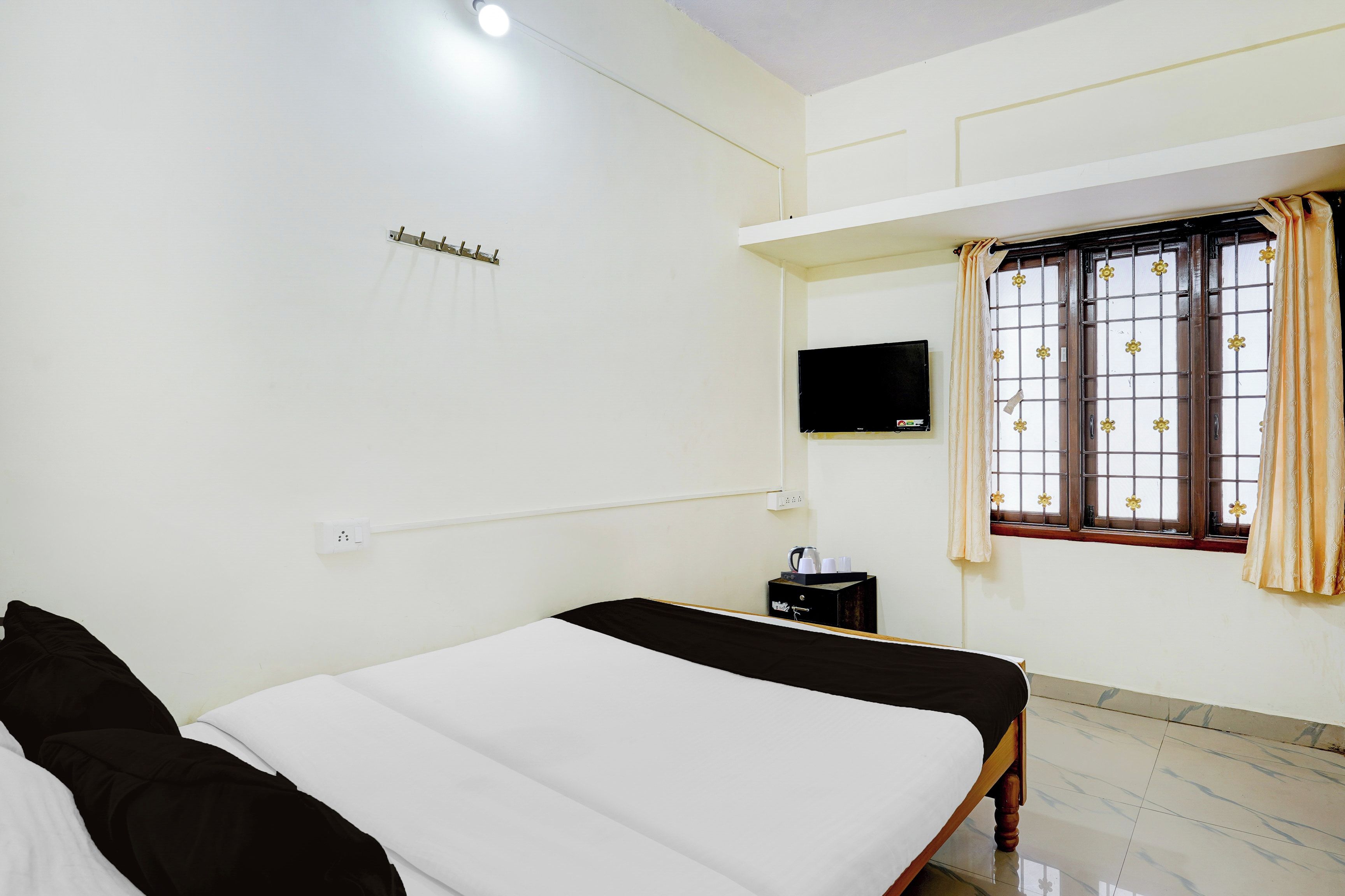 A hotel room with a bed, a television, a window with curtains, and a small table with a kettle and cups.