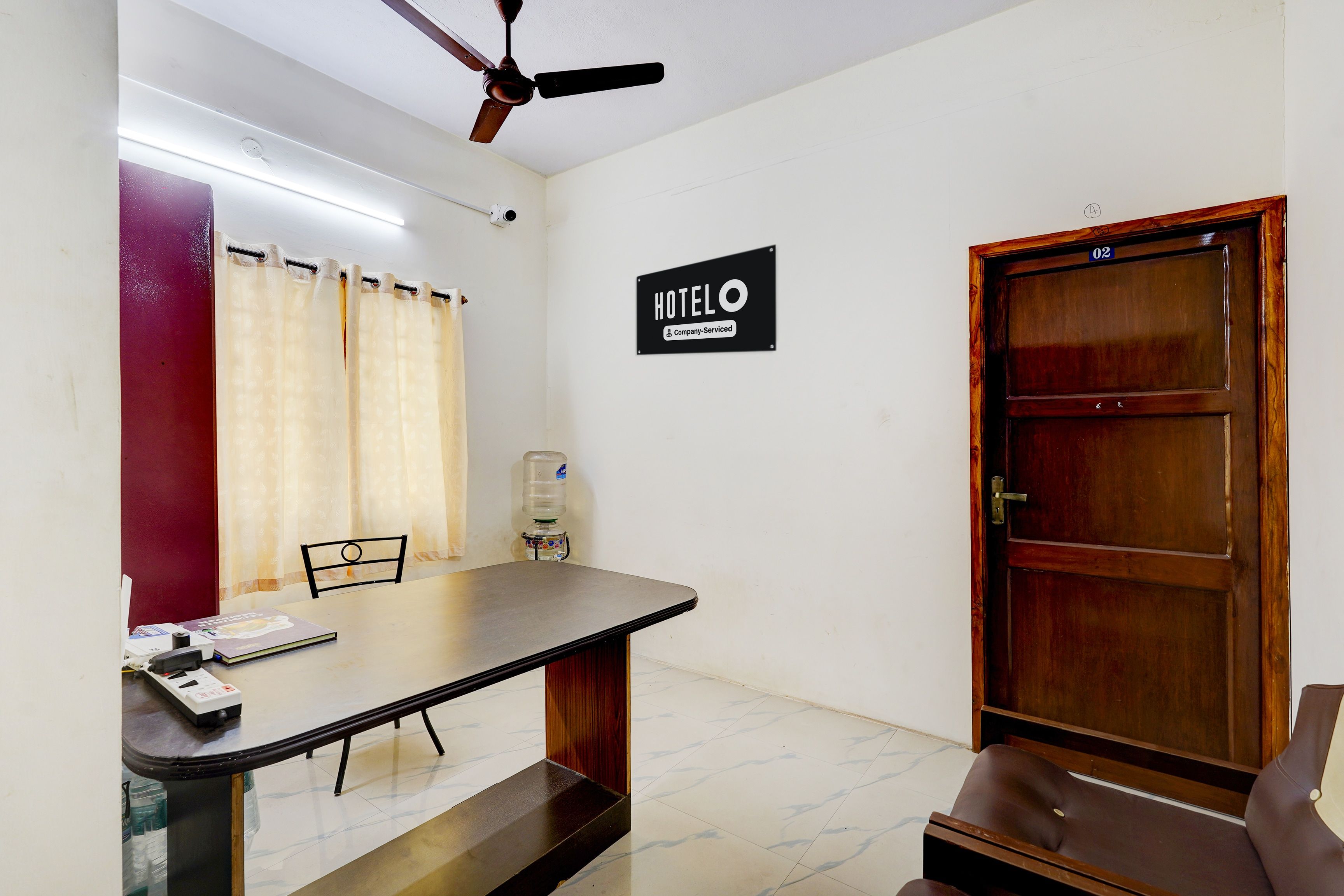 A hotel room with a bed, a desk, a chair, a water dispenser, a ceiling fan, and a door.