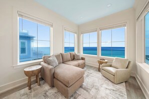 Un salon spacieux comprenant un grand canapé d’angle, un fauteuil et une petite table ronde. La pièce est dotée de grandes fenêtres offrant une vue sur l’océan.