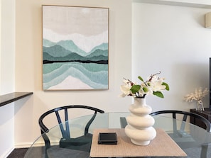 A dining area with a glass table, black chairs, a vase with white flowers, and a framed abstract painting on the wall. - Perth City 2BR Apartment | Pool & Parking (East Perth)