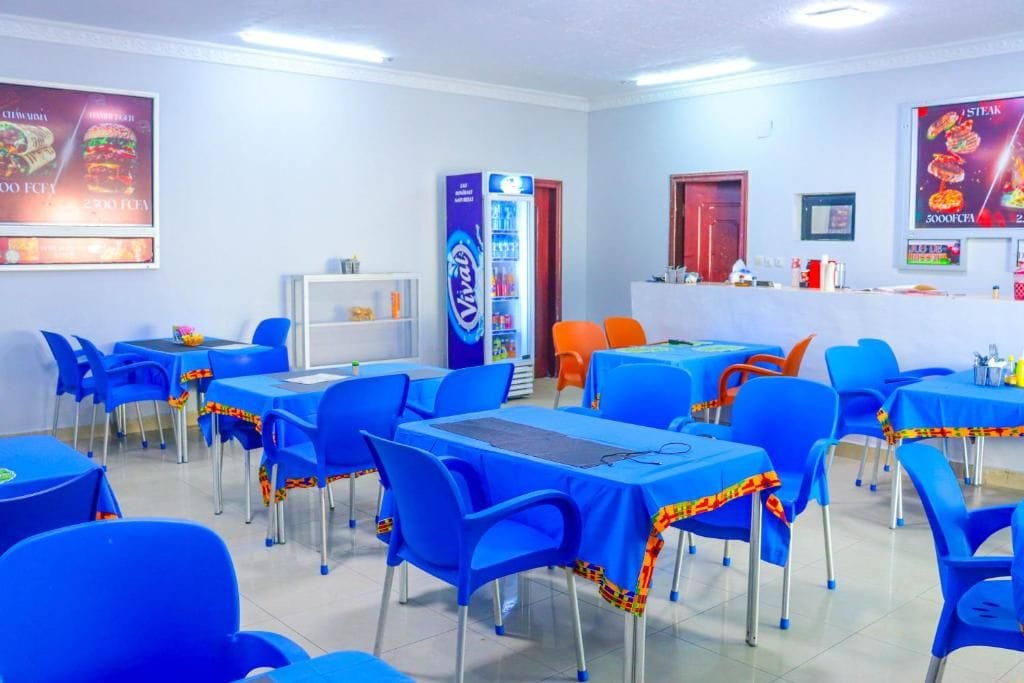 A restaurant with blue tables and chairs, a refrigerator, and a counter with food items.