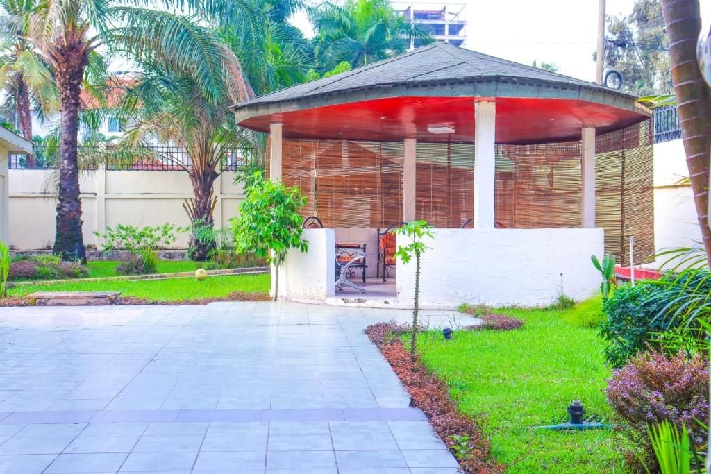A covered outdoor seating area with a bench and a small table, surrounded by a garden with palm trees and shrubs.
