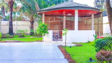 A covered outdoor seating area with a bench and a small table, surrounded by a garden with palm trees and shrubs.