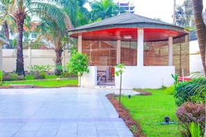 A covered outdoor seating area with a bench and a small table, surrounded by a garden with palm trees and shrubs.