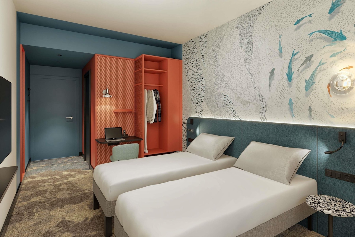 A hotel room with two beds, a desk with a laptop, a wardrobe, and a wall with a fish pattern.
