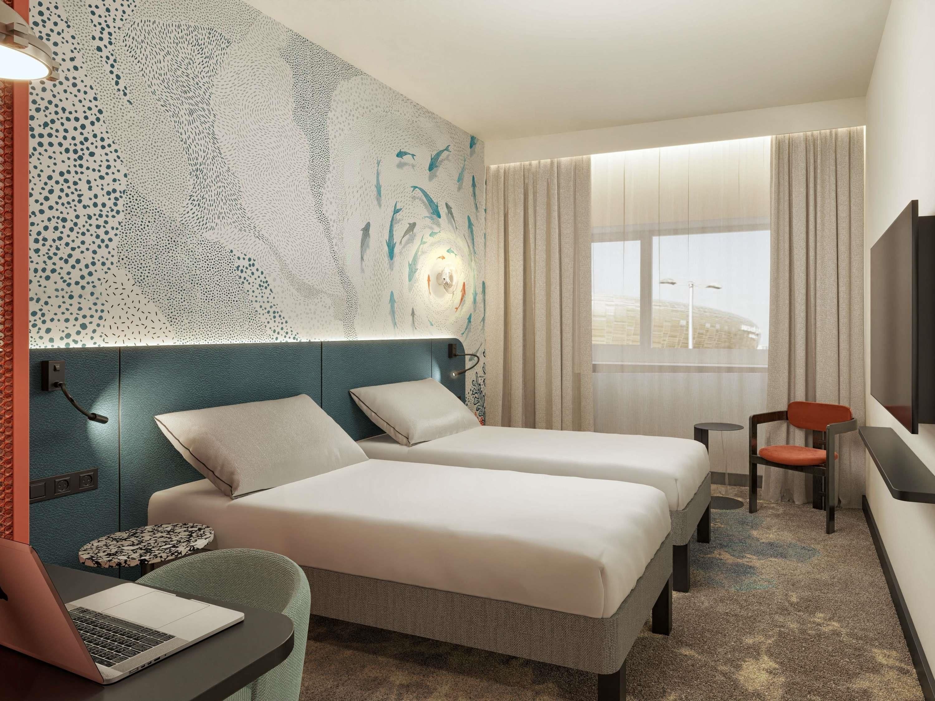 A hotel room with two beds, a desk, a chair, a laptop, and a large window with curtains.