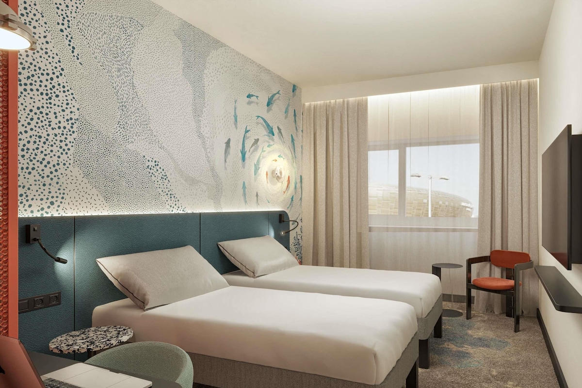 A hotel room with two beds, a desk, a chair, a laptop, and a large window with curtains.