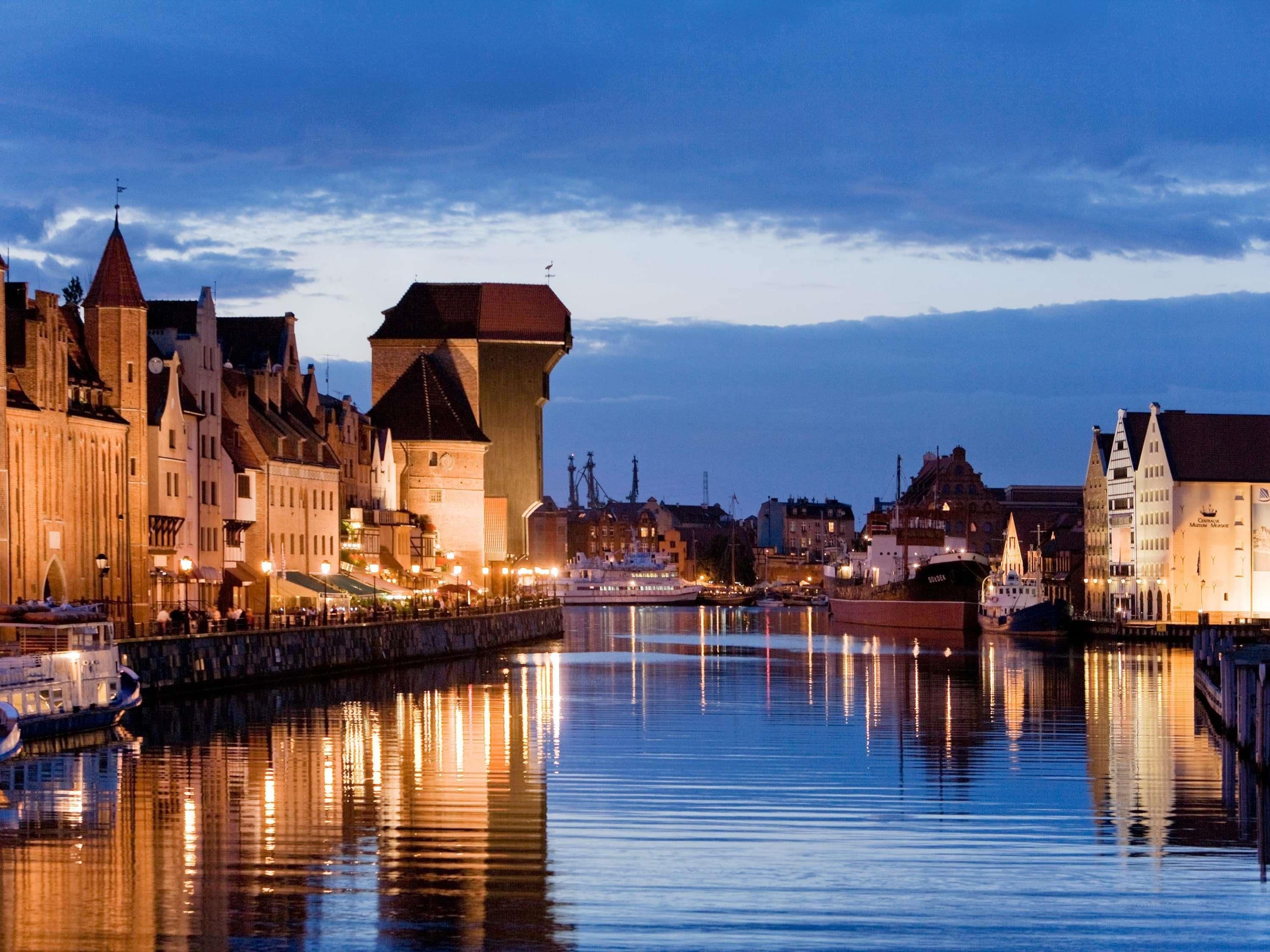 Eine Uferstadt mit historischen Gebäuden, einem markanten Turm und ruhigem Wasser, das die Lichter spiegelt.