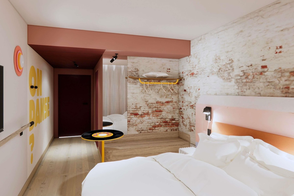 A hotel room with a bed, a small table, a wall with a 'ON DANCE' sign, a brick wall, and a wooden floor.