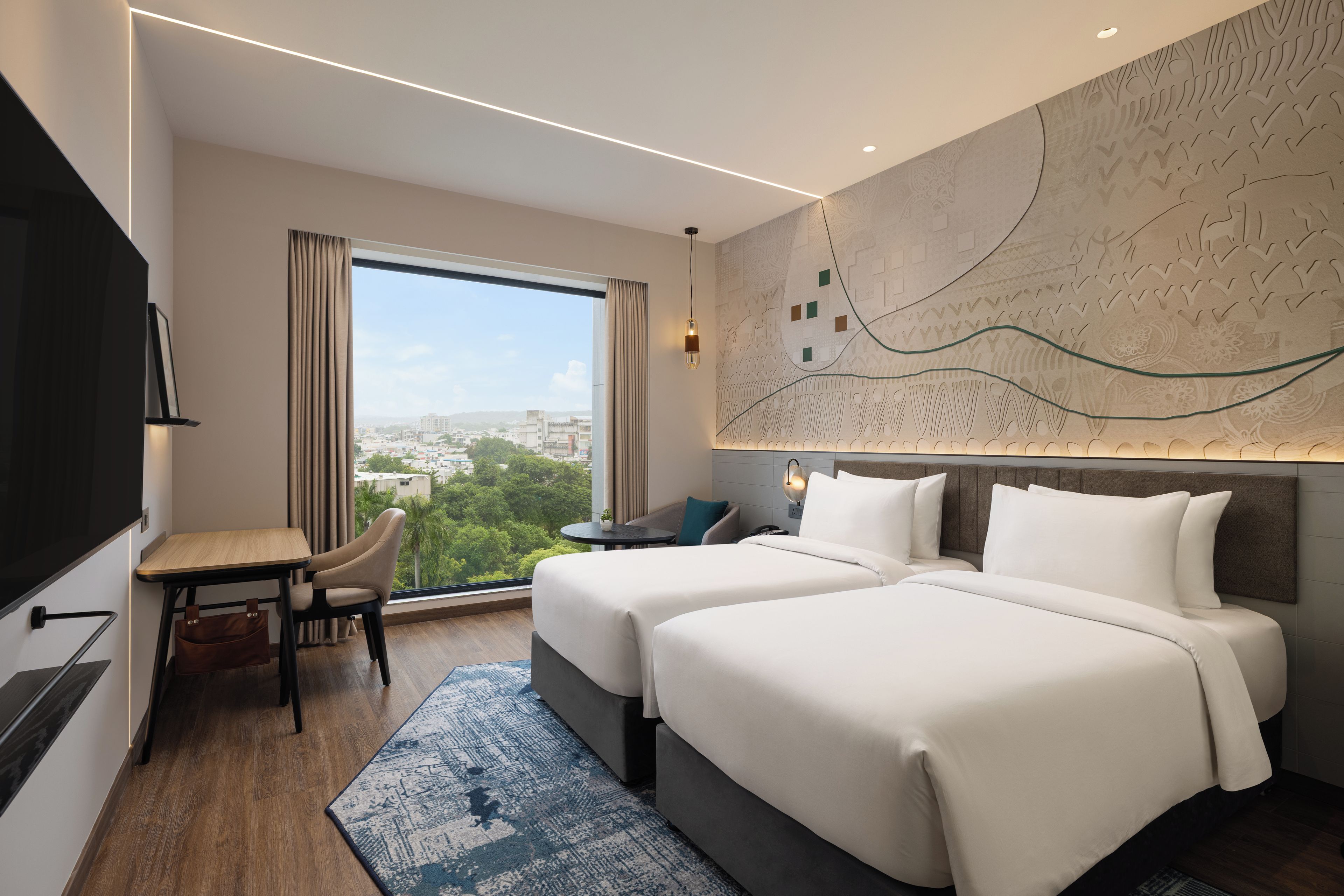 A hotel room with two beds, a desk, a chair, a TV, and a large window offering a city view.