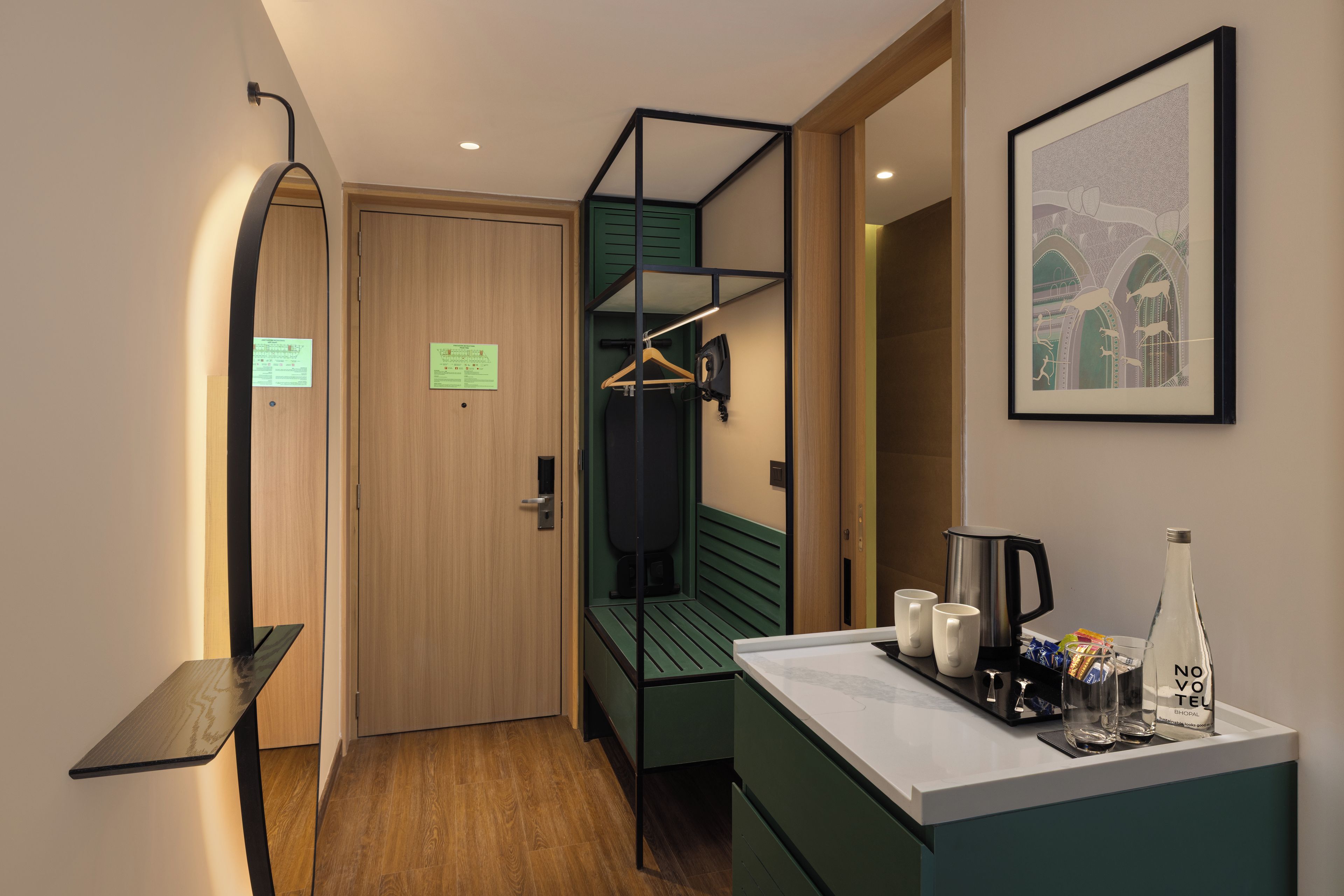 Una habitación de hotel compacta con una puerta de madera, una silla colgante verde, una pequeña kitchenette con cafetera y un cuadro en la pared.