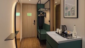 Ett kompakt hotellrum med en trädörr, en grön hängstol, ett litet pentry med en kaffebryggare och en inramad tavla på väggen.