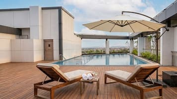Area piscina sul tetto con poltrona prendisole, tavolino con asciugamani e bevande, e un ombrellone grande.