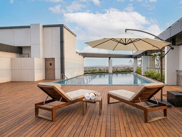 A rooftop pool area with lounge chairs, a table with towels and drinks, and a large umbrella.