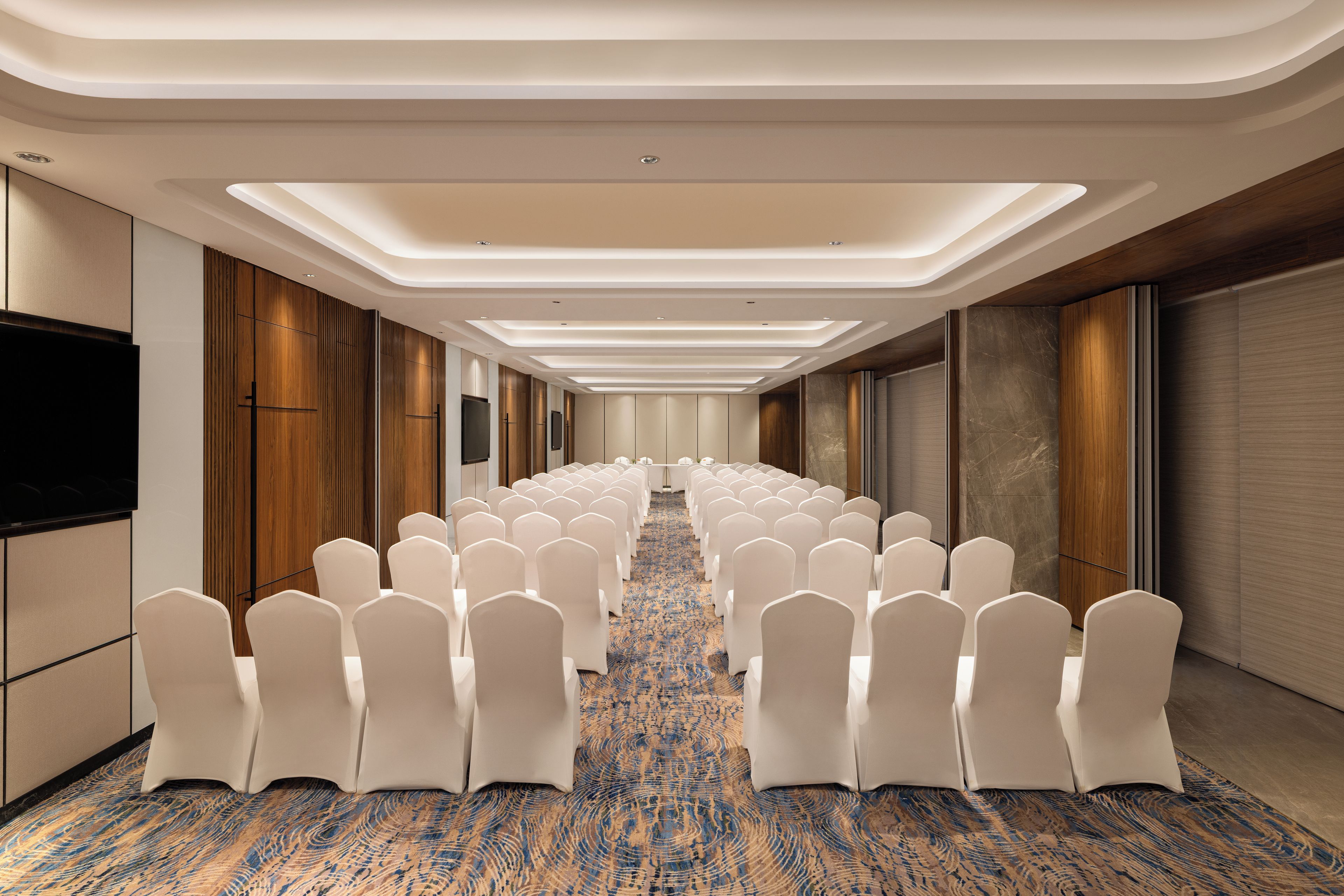 A hotel conference room with rows of white chairs, a patterned carpet, and a modern ceiling design.
