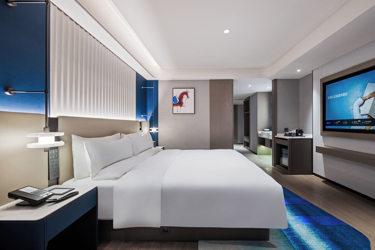 Habitación de hotel moderna con una cama grande, mesita de noche con teléfono y libreta, televisor de pantalla plana en la pared y un cuadro sobre la cama.