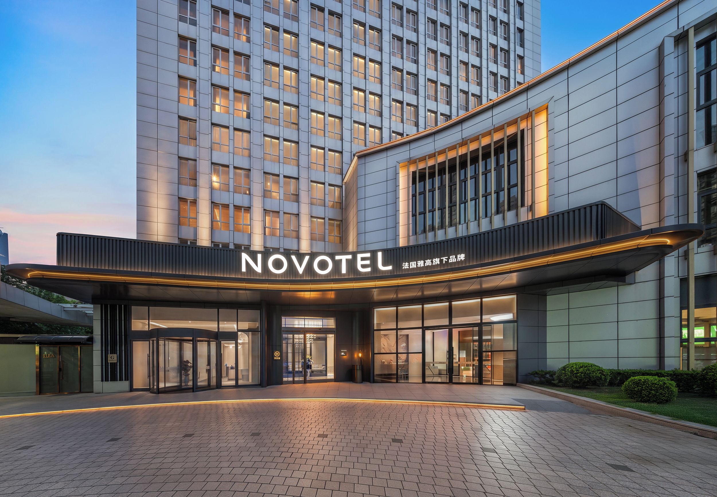 Una entrada de hotel moderna con un llamativo letrero 'NOVOTEL', fachada de vidrio y un camino pavimentado que conduce a la entrada.
