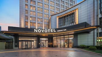 Una entrada de hotel moderna con un llamativo letrero 'NOVOTEL', fachada de vidrio y un camino pavimentado que conduce a la entrada.