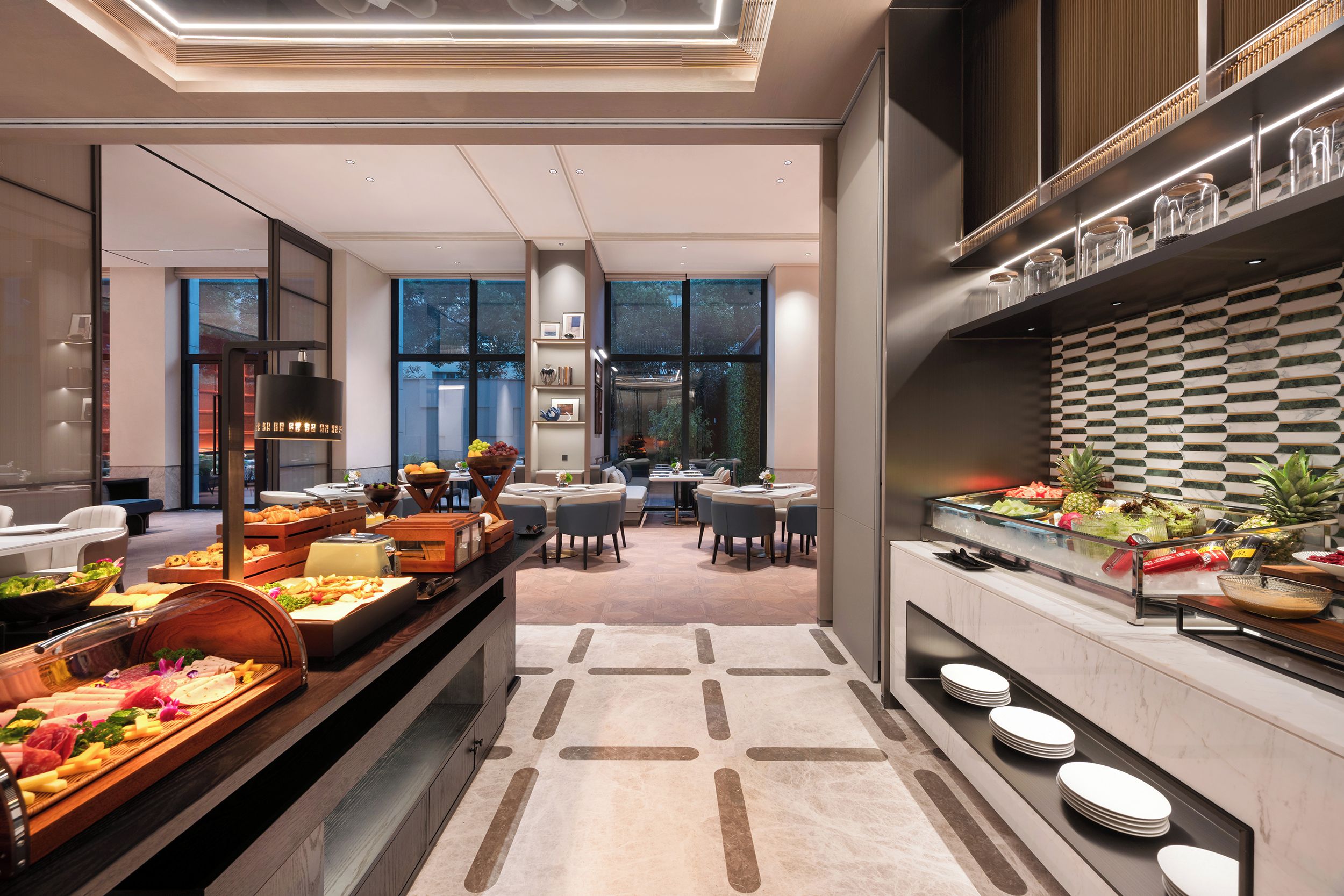 A modern hotel dining area with a buffet setup, seating arrangements, and a view of the outdoors.