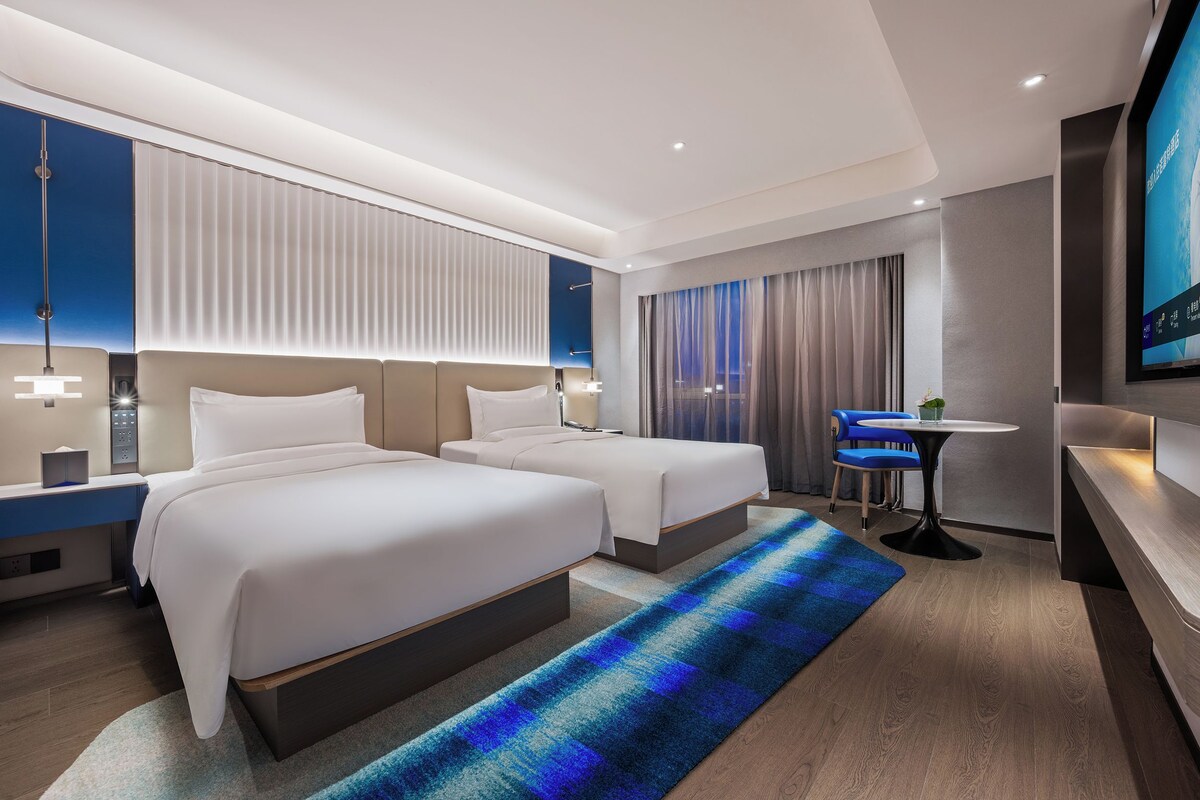 Una habitación de hotel moderna con dos camas, un escritorio, una silla, un televisor y un ventanal con cortinas.