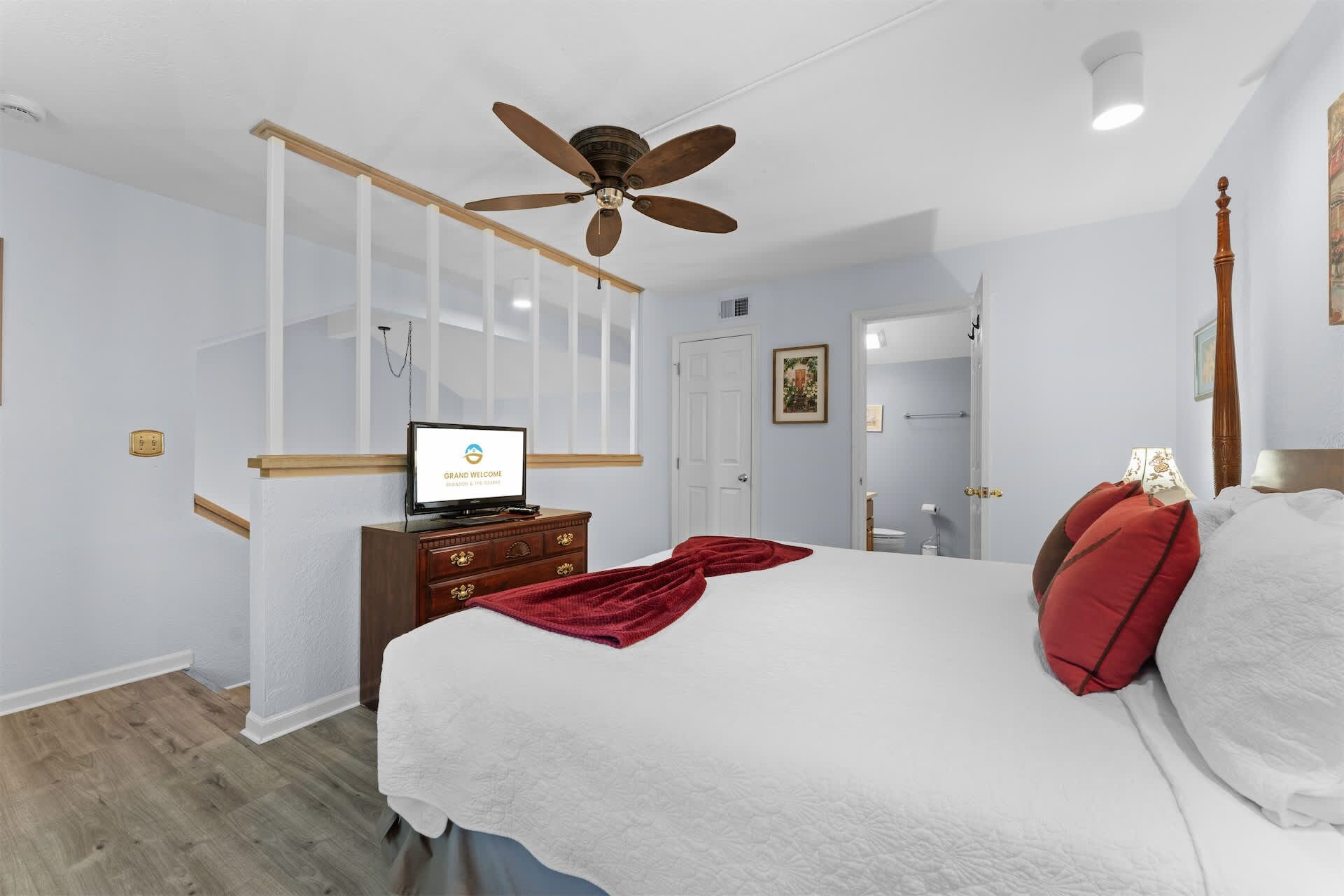 A hotel room with a bed, a ceiling fan, a TV on a dresser, and a door leading to a bathroom.