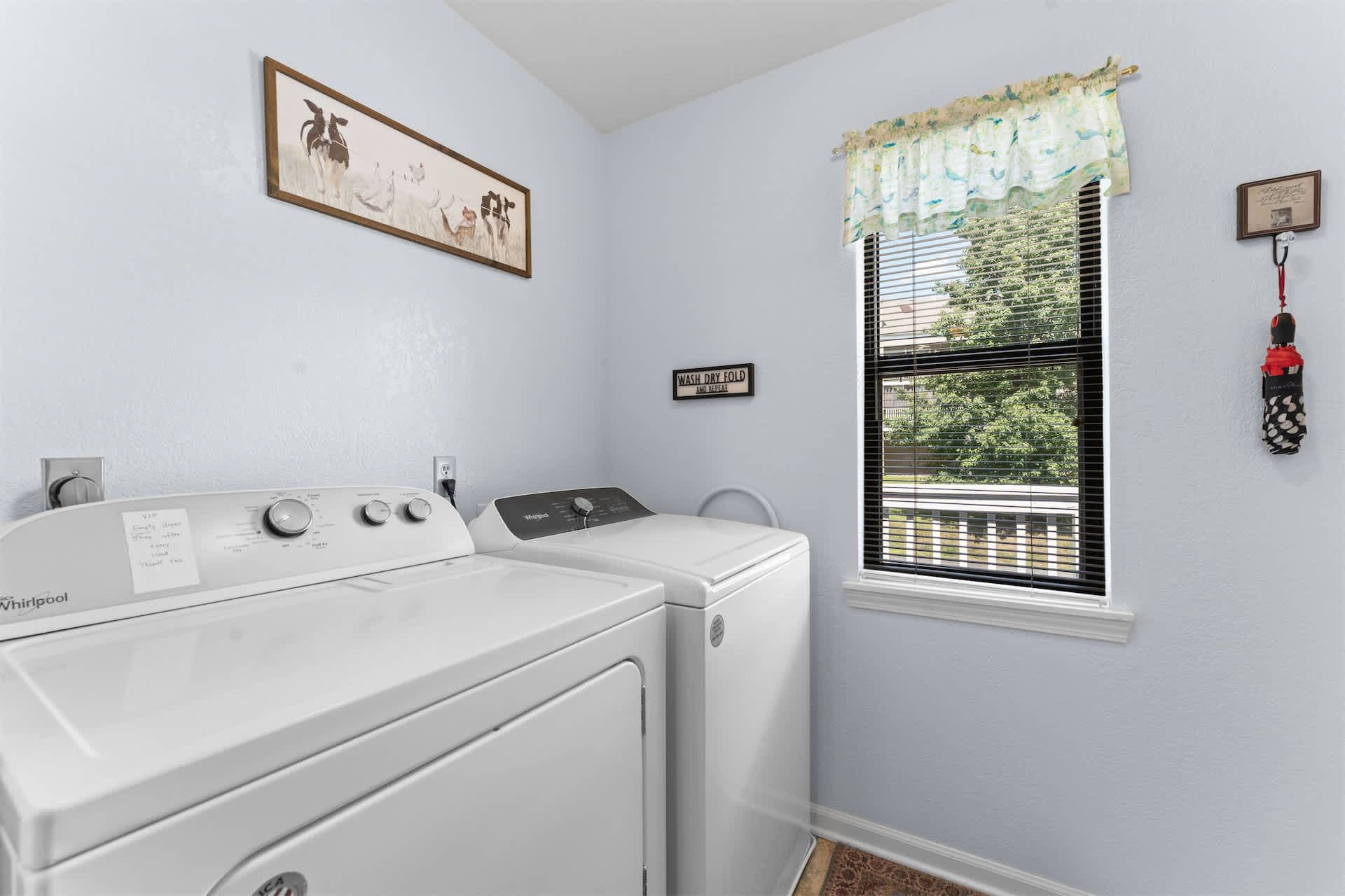 A laundry room with a front-loading washer and dryer, a window with blinds, a framed picture on the wall, and a sign above the dryer.