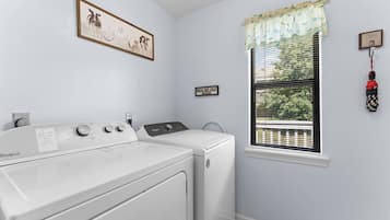 A laundry room with a front-loading washer and dryer, a window with blinds, a framed picture on the wall, and a sign above the dryer.