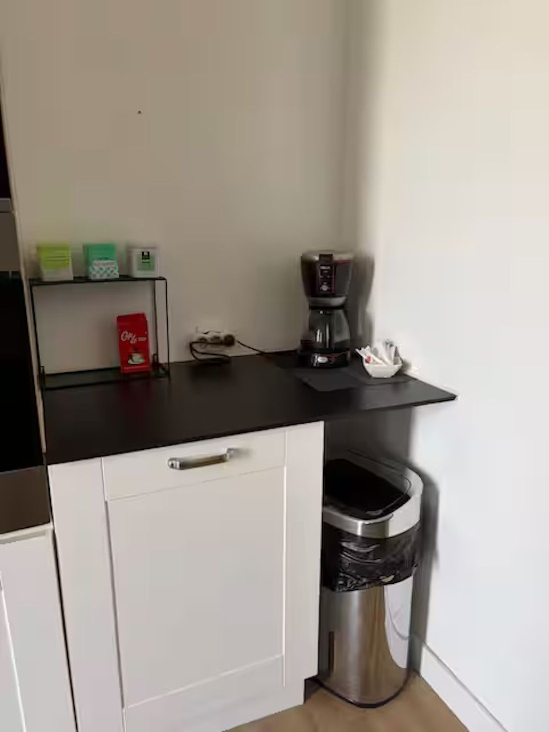 A kitchenette with a coffee maker, a trash bin, and a shelf with items.