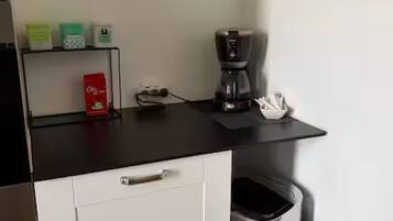 A kitchenette with a coffee maker, a trash bin, and a shelf with items.