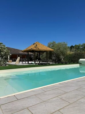 A swimming pool with a thatched-roof pavilion, a stone wall, and a grassy area. - Le Pignole · 2-bedroom apartment with AC and parking · Shared pool and garden · Sleeps 5 (Saint-Jory)