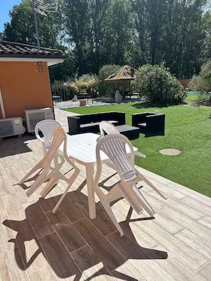 An outdoor patio with a table and chairs, a small building, and a well-maintained lawn. - Le Pignole · 2-bedroom apartment with AC and parking · Shared pool and garden · Sleeps 5 (Saint-Jory)