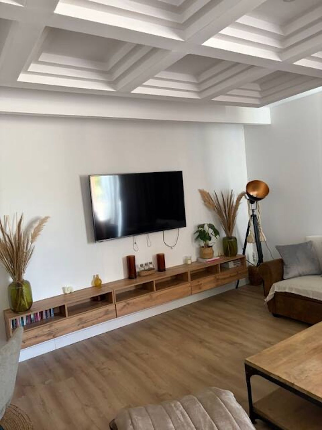 Een moderne woonkamer met een flatscreen televisie aan de muur, houten meubels en een plafond met geometrische patronen.