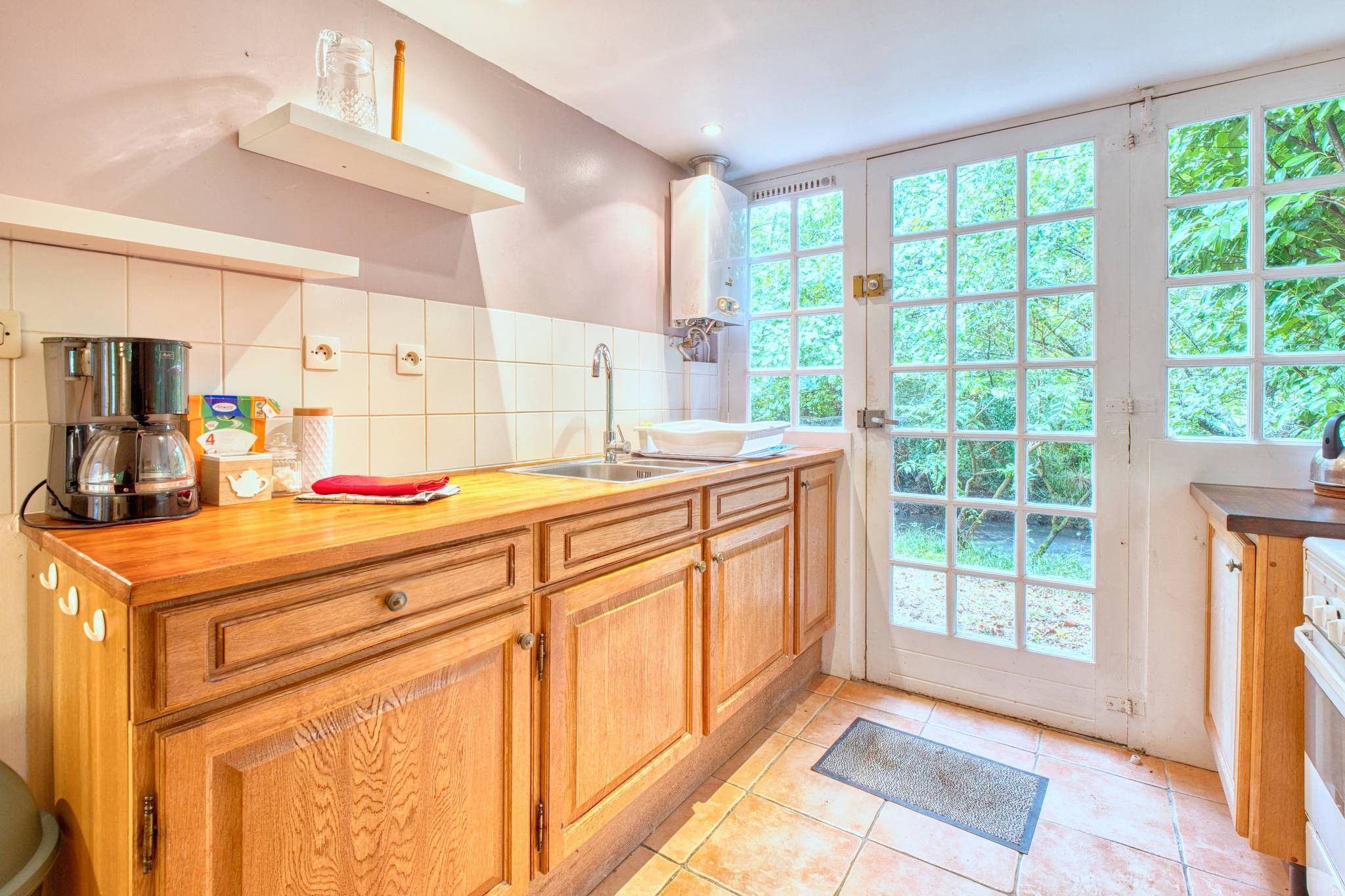 A kitchen with wooden cabinets, a coffee maker, a sink, and a window with a view of greenery.