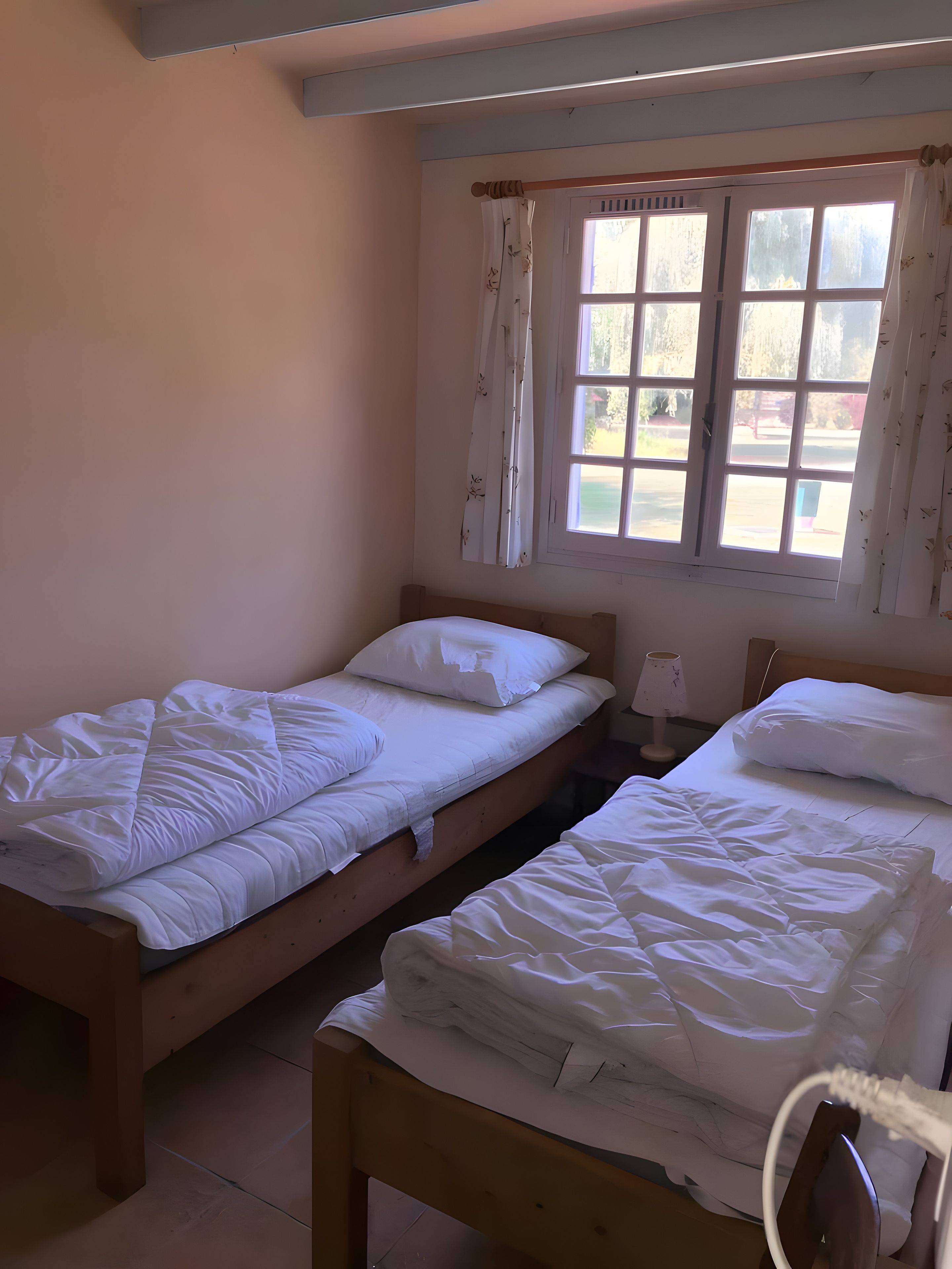A room with two single beds, a window with curtains, a bedside table with a lamp, and a wall-mounted shelf.