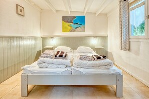 Un letto rifatto con coperte e cuscini piegati, un quadro sopra e una finestra con tende.