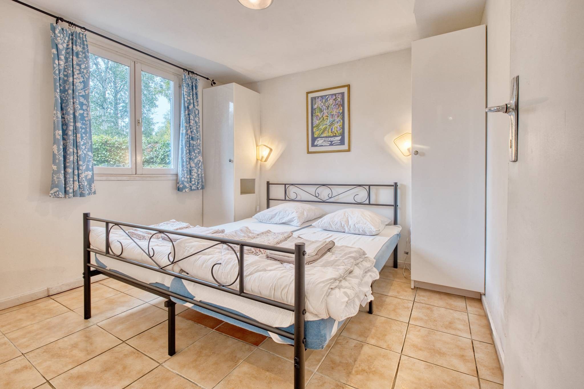 A bedroom with a metal bed frame, white bedding, a window with curtains, a wall-mounted light, and a framed picture.