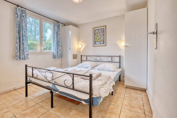 Un dormitorio con estructura de cama metálica, ropa de cama blanca, una ventana con cortinas, una lámpara fijada a la pared y un cuadro enmarcado.