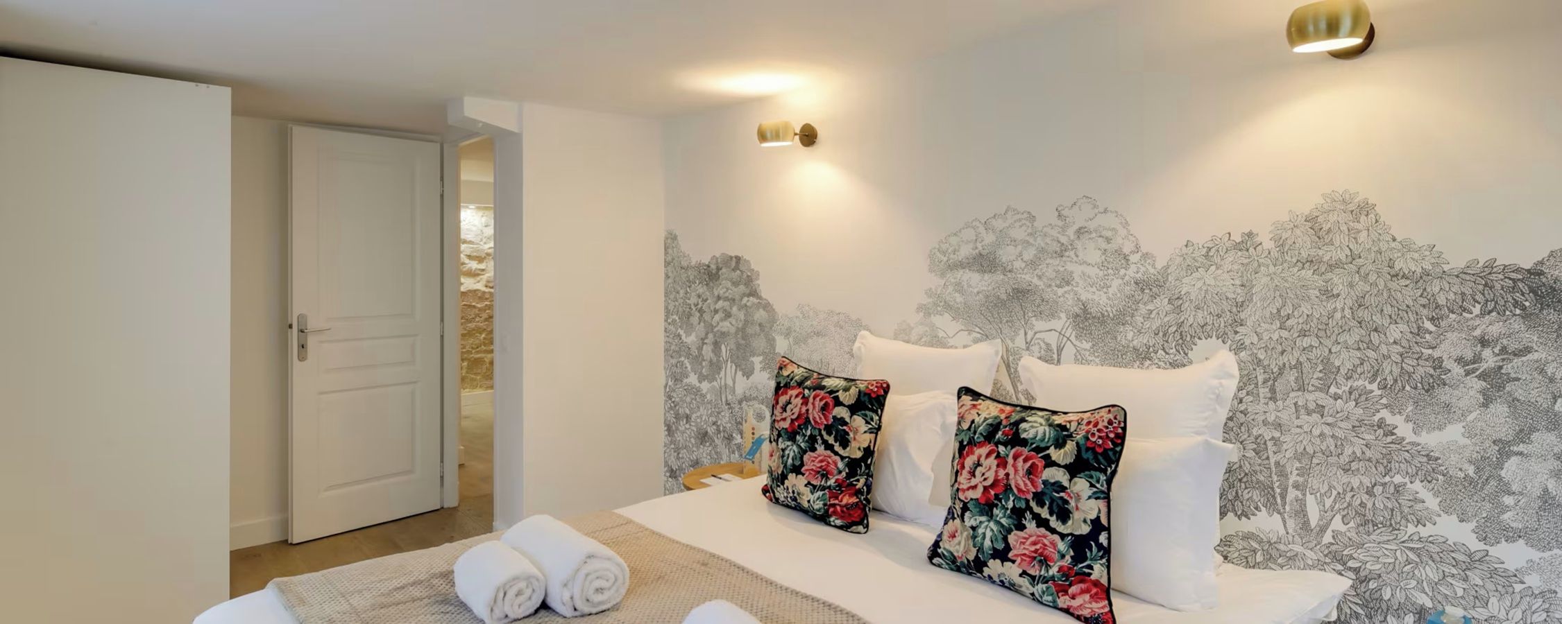 A hotel room with a bed, pillows, a wall with a floral pattern, a door, and a light fixture.