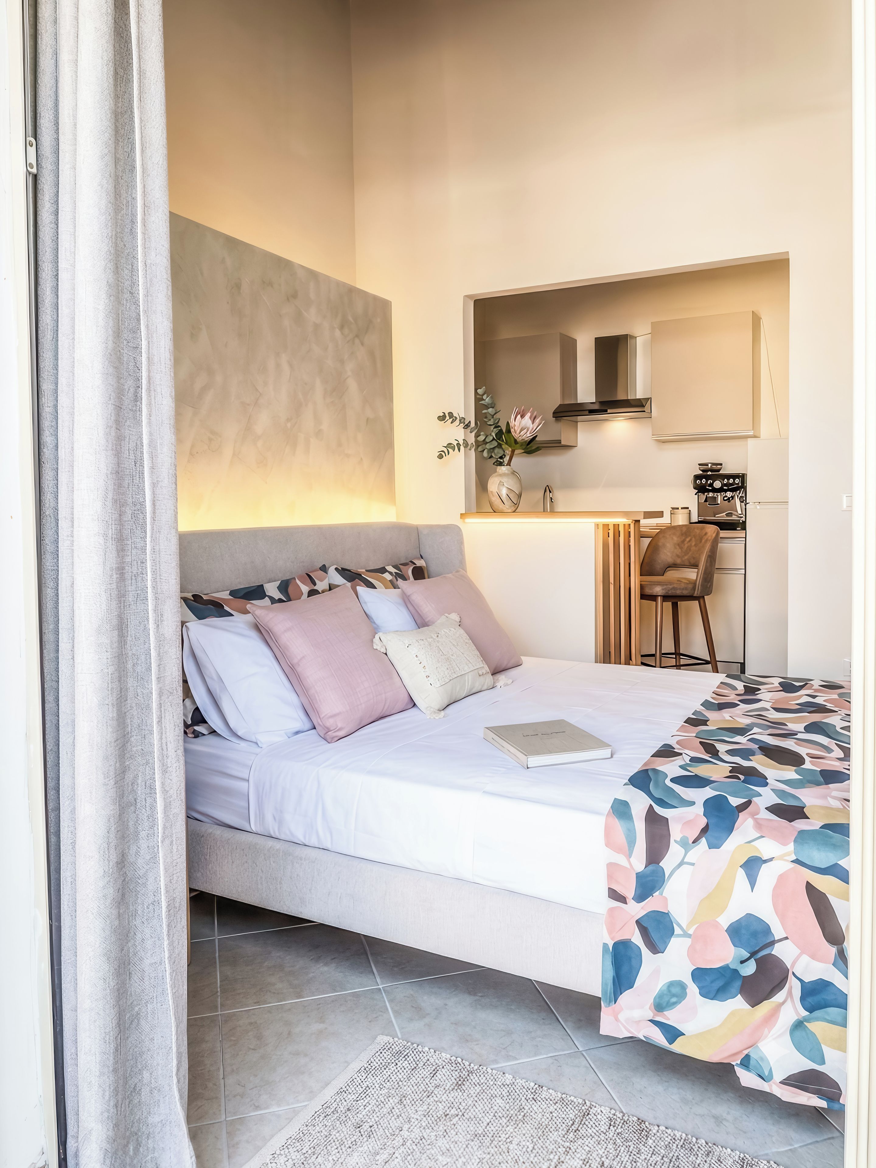 A bedroom with a bed, a bedside table, a chair, a small kitchenette, and a bathroom visible through an open door.