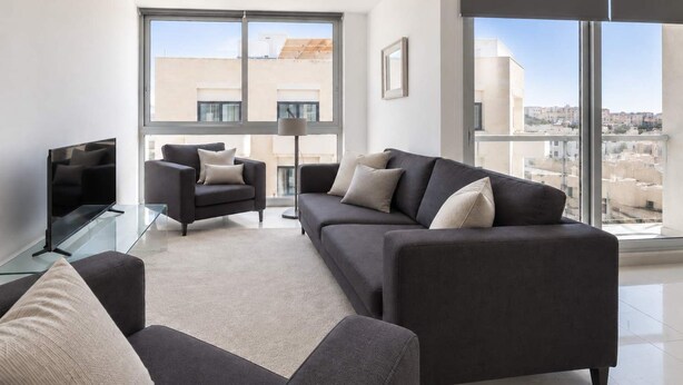 En moderne stue med en stor sofa, en mindre lænestol, et sofabord i glas og et fladskærms-tv.