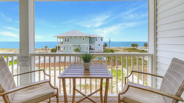 Eine überdachte Veranda mit Blick auf einen Strand und ein Haus. Ein Tisch mit einer Topfpflanze und zwei Korbstühle stehen bereit. Ein Warnschild ist am Geländer angebracht.