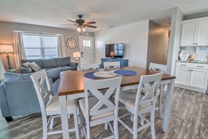 A dining area with a wooden table and white chairs, a television in the background, and a kitchen with white cabinets. - BEACON BREAK PAD IS 350 STEPS TO BEACH! 4BD/2BA, PRIVATE POOL! (Port Saint Joe)